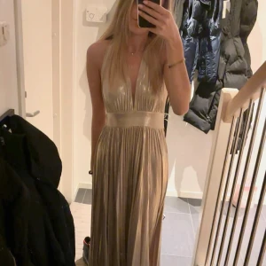 Guldig långklänning med öppen rygg - Elegant guldig långklänning med djup v-ringning och öppen rygg. Klänningen har en knytning i nacken och ett brett band i midjan som ger en smickrande passform. Perfekt balklänning! Storlek xxs men borde passa xs också✨✨ Klänningen har aldrig använts, bara provats en gång. Säljer eftersom den var något för stor.