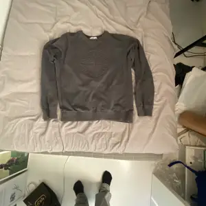 Säljer en stilren grå sweatshirt från Stone Island med ett diskret broderat logotypmärke på bröstet. Tröjan har långa ärmar och en rund halsringning med ribbade muddar vid ärmslut och nederkant. Perfekt för en avslappnad look.