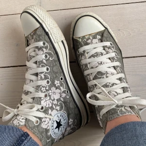 Blommiga Converse All Star sneakers - Coola Converse All Star sneakers i den höga modellen med ljusrosa blomsterslingor på en grå bakgrund med dödskallar.  Storlek 37,5 men funkar bra på mig som har breda fötter storlek 38. 