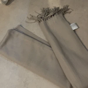 Beige halsduk  - Snygg beige halsduk med fransar i ändarna. Perfekt för att hålla dig varm under kyliga dagar. Enkel och stilren design som passar till det mesta.