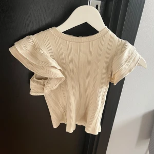 Beige ribbad topp från Gina - Säljer en söt beige ribbad topp med volangdetaljer på ärmarna. Toppen har en kortärmad design och är perfekt för en stilren look. Passar bra till både jeans och kjol.