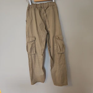 Beige cargobyxor från Lindex - Säljer ett par beige cargobyxor från Lindex med praktiska fickor på sidorna. Byxorna har en knapp och dragkedja framtill samt elastisk midja för extra komfort. Perfekta för en avslappnad stil.