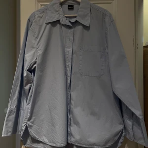 Ljusblå skjorta från Gina Tricot - Snygg ljusblå skjorta från Gina Tricot med klassisk krage och en bröstficka. Skjortan har långa ärmar och en oversized passform. Perfekt för en stilren look.