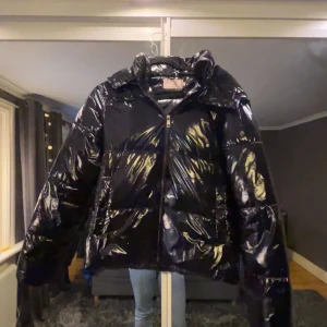 Svart trendig pufferjacka - Säljer en svart trendig glansig pufferjacka med dragkedja och huva. Jackan har en modern look och är perfekt för kyligare dagar. Den är långärmad och har en stilren design som passar till många olika outfits.