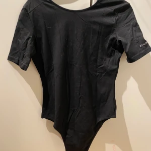 Svart bodysuit från Workout Empire - Säljer en svart bodysuit från Workout Empire med korta ärmar och en djup v-ringning med metallringar på ryggen. Perfekt för träning eller en stilren look. Texten 'WITH CONFIDENCE' finns på ärmen. 