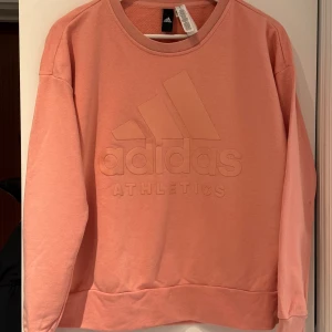 Rosa/Persikofärgad sweatshirt från Adidas - Säljer en snygg rosa/persikofärgad sweatshirt från Adidas Athletics. Tröjan har en stor logga på framsidan och långa ärmar. Perfekt för en sportig och avslappnad stil. Passar bra till både jeans och träningsbyxor.