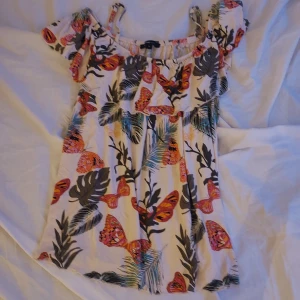 Flerfärgad offshoulder klänning från Wear Wom - Säljer en somrig offshoulder topp från Wear Women med ett färgglatt mönster av fjärilar och blad. Toppen har korta ärmar och en luftig passform, perfekt för varma dagar. Den är i en härlig mix av färger som rosa, orange och grön.