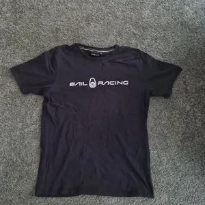 Svart t-shirt från Sail Racing - Snygg svart t-shirt från Sail Racing med tryckt logga på framsidan. Perfekt för en avslappnad stil. Tillverkad i mjukt material för extra komfort.