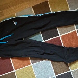 Svarta mjukisbyxor från Emporio Armani - Snygga svarta mjukisbyxor från Emporio Armani med blå detaljer längs sidorna. Perfekta för en avslappnad stil. Byxorna har en bekväm passform och är gjorda i mjukt material.