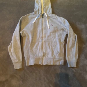 Grå hoodie från Polo Ralph Lauren - Säljer en stilren grå hoodie från Polo Ralph Lauren med dragkedja och justerbar huva. Den har en liten broderad logga på bröstet och praktiska fickor framtill. Perfekt för en avslappnad look.