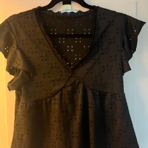 Svart blus från Shein - Säljer en svart blus från Shein med v-ringning och spetsdetaljer. Blusen har korta ärmar och ett fint broderat mönster. Perfekt för en stilren look.