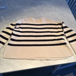 Randig stickad tröja från GAP Kids - Säljer en snygg randig stickad tröja från GAP Kids i beige och svart. Tröjan har tre fjärdedels arm  och en klassisk rund halsringning. Perfekt för en stilren look! Den har en liten fläck på framsidan men man märker knappt av den. Barn-Medium