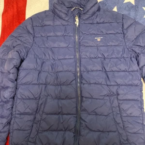 Blå pufferjacka från GANT - Säljer en snygg blå pufferjacka från GANT. Jackan har en dragkedja framtill och är quiltad för extra värme. Perfekt för kyligare dagar. Den har en stilren design med GANT-logga på bröstet.