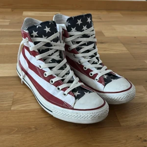 Converse All Star med amerikanskt flaggmönster - Snygga Converse All Star sneakers med ett iögonfallande amerikanskt flaggmönster. Skorna har klassisk snörning och hög modell med stjärnor och ränder i rött, vitt och blått. Perfekta för att sticka ut i mängden!