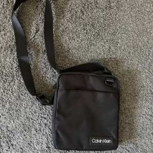 Svart axelväska från Calvin Klein - Snygg och praktisk svart axelväska från Calvin Klein. Väskan har en justerbar axelrem och en dragkedja för säker förvaring. Perfekt för att bära med sig det nödvändigaste med stil. Nyskick