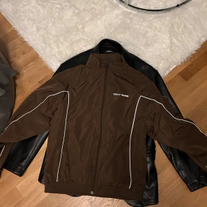 Brun vindjacka från Daily Paper - Snygg brun vindjacka från Daily Paper med dragkedja och vita detaljer längs ärmarna. Perfekt för en stilren look. Jackan har en hög krage och normal passform.