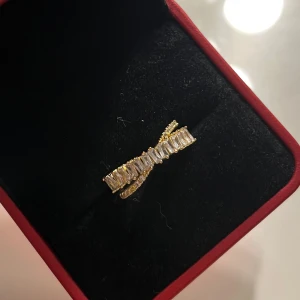 Guldfärgad ring med glittriga stenar - Elegant guldfärgad ring med en rad av glittriga stenar i en korsande design. Perfekt för att lägga till en touch av glamour till din stil. 