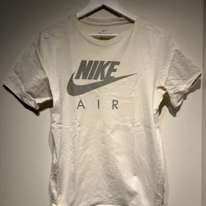 Vit t-shirt från Nike - Säljer en vit t-shirt från Nike med det klassiska Nike Air-logotypen i grått på framsidan. T-shirten har en rund hals och korta ärmar, perfekt för en avslappnad stil.