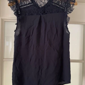 Blå blus med spetsdetaljer - Elegant blå blus med vackra spetsdetaljer vid axlarna och övre delen. Blusen har knappar baktill och är perfekt för en stilren look.  Alla lappar är bortklippta. 