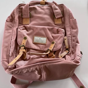 Rosa ryggsäck från Doughnut - Säljer en superfin rosa ryggsäck från Doughnut. Den har snygga läderdetaljer och en praktisk ficka framtill med spännen. Perfekt för skolan eller en dag på stan! 💖