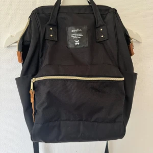 Svart ryggsäck från Anello - Säljer en snygg och praktisk svart ryggsäck från Anello. Den har en stilren design med dragkedjor i guld och bruna detaljer. Ryggsäcken är rymlig med flera fack, perfekt för både skola och vardag. Den är i ny skick och redo för nya äventyr!