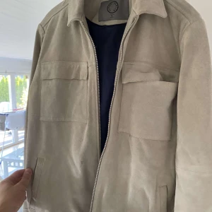 Beige mockajacka med dragkedja - Säljer en snygg beige mockajacka med dragkedja framtill. Jackan har två stora bröstfickor och en stilren design. Perfekt för höst och vår, och den är långärmad för extra värme. Passar både till vardags och lite finare tillfällen.