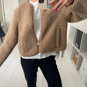 Beige teddyjacka med dragkedja - Säljer en supermysig beige teddyjacka som är perfekt för höst och vinter. Den har en dragkedja framtill och är långärmad. Jackan är croppad och ger en avslappnad look. Perfekt för att hålla sig varm och stilren samtidigt!