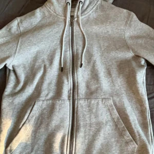 Grå hoodie från Jack & Jones - Säljer en snygg och bekväm grå hoodie från Jack & Jones. Den har en dragkedja framtill och en praktisk ficka på magen. Perfekt för en avslappnad stil och passar till alla tillfällen. Huvan har dragsko för justering. Perfekt för höst och vår!