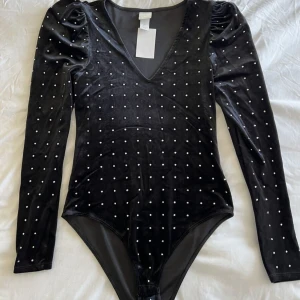 Svart sammetstopp med prickar - Säljer en elegant svart body i sammet med små prickar över hela tyget. Toppen har en djup v-ringning och långa ärmar med lätt puff vid axlarna. Perfekt för en festlig kväll eller en utekväll med vänner!