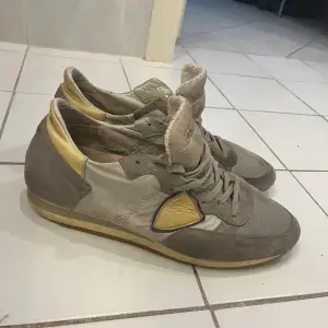 Säljer ett par grå och beige sneakers från Philippe Model. De har en snygg kombination av mocka och textilmaterial, vilket ger dem en unik look. Skorna har snörning och en bekväm passform, perfekt för vardagsbruk. De är i välanvänt skick men har fortfarande mycket liv kvar.