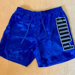 Blå Puma shorts stl XL - Sköna Puma shorts i glansigt material, stl XL. Bra skick, endast små tecken på användning. Stor tryckt logga och en broderad logga på benen.