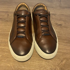 Bruna Boss Skinn Skor - Säljer ett par snygga bruna sneakers från Boss i skinn. De har en stilren design med snörning och en vit sula som ger en fräsch kontrast. Storlek 44, mildt använda och i mycket välomhändertaget skick! Nypris: 4799, mitt pris är öppen till förhandling!