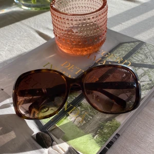 Solglasögon från Prada - Säljer ett par snygga solglasögon från Prada. De har en klassisk brun färg med ett stilrent mönster som påminner om sköldpaddsskal. Glasögonen har en rektangulär form och är perfekta för soliga dagar. De är i bra skick och har en elegant design med Pradas logga på sidan. Perfekta för att ge din outfit en lyxig touch!