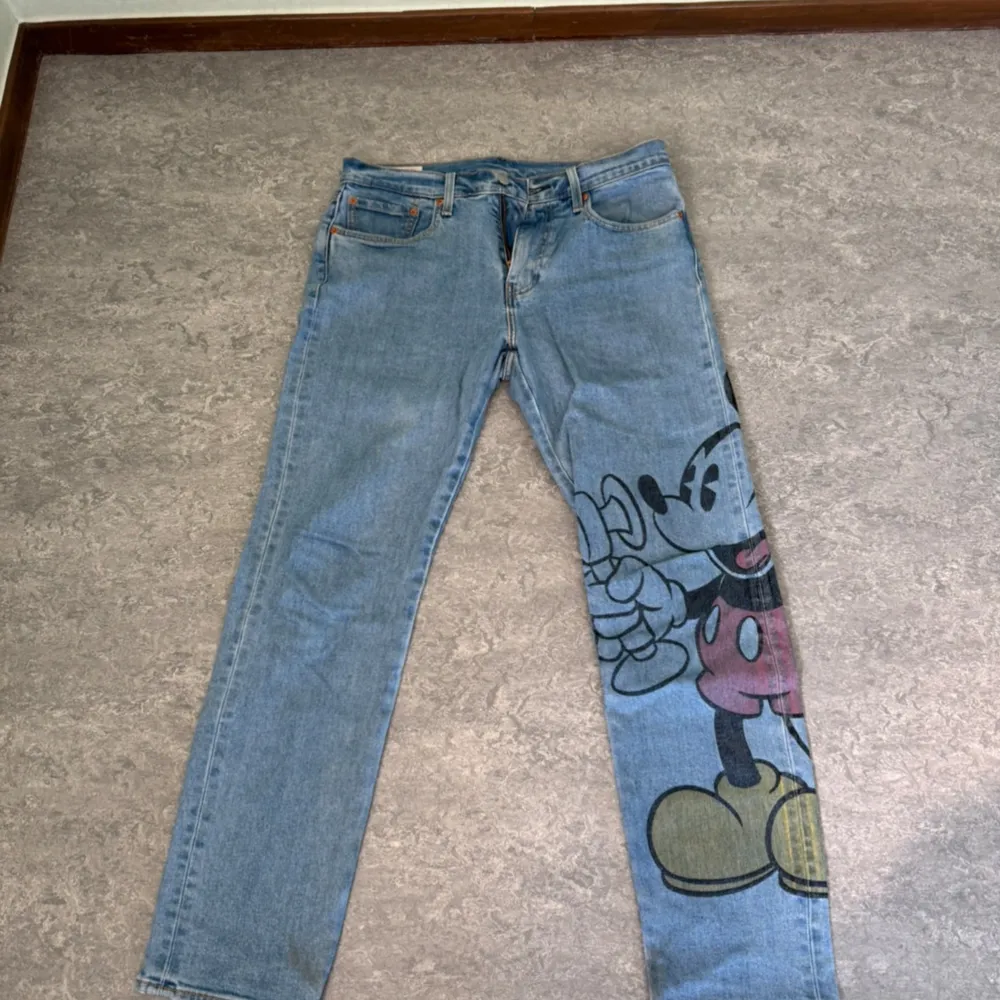 Säljer ett par coola jeans från Levis (502-modell) med ett stort Musse Pigg-tryck på ena benet. De är ljusblå och har en klassisk femficksdesign. Perfekta för dig som vill sticka ut lite extra! 🐭👖. Jeans & Byxor.