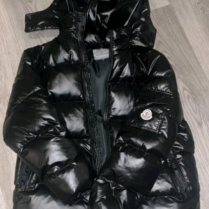 Svart pufferjacka från Moncler - Säljer en snygg och varm svart pufferjacka från Moncler. Jackan har en glansig finish och är perfekt för kalla vinterdagar. Den har en dragkedja framtill och en praktisk huva. Insidan är fodrad för extra värme och komfort. Perfekt för dig som vill hålla stilen även när det är kallt ute!