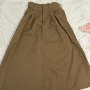 Beige kjol med hög midja - Säljer en superfin beige kjol med hög midja. Den har en stilren design med veckdetaljer upptill och en elastisk midja för extra komfort. Perfekt för både vardag och festliga tillfällen!