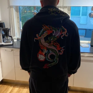 Svart Ed Hardy hoodie med drakmotiv - Säljer en svart hoodie från Ed Hardy med ett coolt drakmotiv på ryggen. Den har en dragkedja framtill och en huva med snörning. På ärmen och bröstet finns Ed Hardy-loggan i gult. Perfekt för en avslappnad och stilren look!