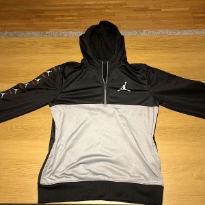 Svart och grå hoodie från Jordan - Säljer en snygg svart och grå hoodie från Jordan. Den har en halv dragkedja framtill och en stor logga på bröstet. På ärmarna finns det coola detaljer med Jordan-loggan och texten 'Flight'. Perfekt för både träning och vardag!