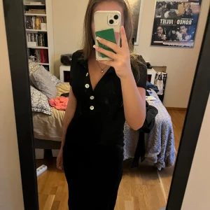 Svart klänning med knappar - Säljer en elegant svart klänning med knappar framtill. Den har korta ärmar och en v-ringad design med krage. 