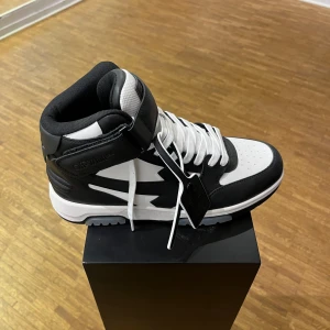 Svartvita sneakers med snörning - Sprillans ny OFF-WHITE sneakers med hög ankel och snörning. De har en cool design med en bred rem över vristen för extra stil och stöd. Perfekta för både vardag och fest!