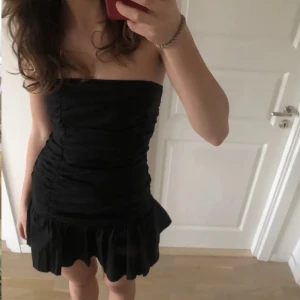 Svart axelbandslös klänning - Säljer en snygg svart axelbandslös klänning med volangdetaljer längst ner. Klänningen är tight och perfekt för festliga tillfällen. Liknar klänningen ifrån Maje!💕