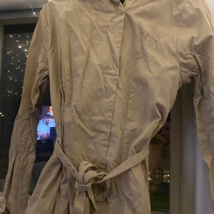 Beige trenchcoat från Bik Bok - Säljer en snygg beige trenchcoat från Bik Bok i storlek M. Den har ett bälte i midjan för en fin passform och är perfekt för vår och höst. Jackan är långärmad och har en stilren design. Perfekt för att ge din outfit en klassisk touch!