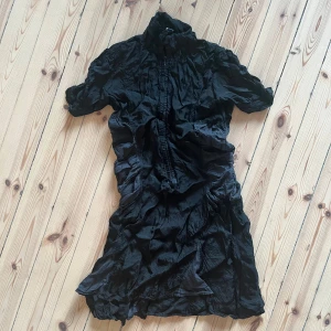 Svart klänning med korta ärmar - Säljer en svart klänning med korta ärmar. Den har en unik rynkad design och knappar framtill. Perfekt för en festlig kväll eller en stilren vardagslook. Jean Paul Gaultier. Lite trasig i ryggen tyvärr (kan skicka fler bilder) därav billig