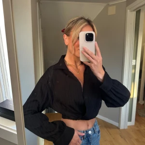 Svart croppad skjorta - Säljer en snygg svart croppad skjorta med långa ärmar. Perfekt för en stilren look, oavsett om det är till vardags eller fest. Skjortan har en klassisk krage och är i ett mjukt material som känns skönt mot huden.