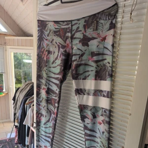 Mönstrade leggings i polyester och spandex - Säljer ett par snygga leggings med tropiskt mönster i grönt, rosa och svart. De har en bred vit midja och två vita ränder på ena benet. Materialet är 90% polyester och 10% spandex, vilket gör dem stretchiga och bekväma. Perfekta för träning eller en avslappnad dag!
