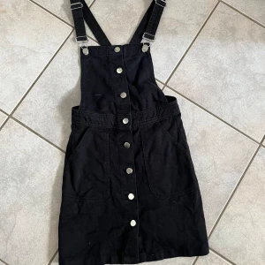 Svart hängselklänning från Divided - Säljer en superfin svart hängselklänning från Divided. Den har justerbara axelband och knappar framtill som ger en cool look. Perfekt för både vardag och fest! 🖤 skriv vid frågor💗