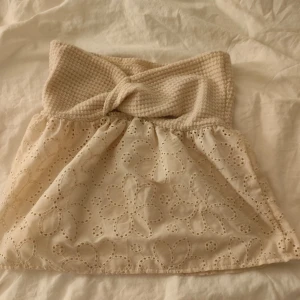 Beige axelbandslös topp med spetsdetaljer - Säljer en superfin beige axelbandslös topp med en stickad överdel och spetsdetaljer på nederdelen. Perfekt för sommardagar eller en festlig kväll. Toppen har en knutdetalj framtill som ger en extra touch av stil.