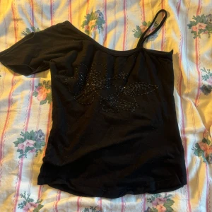 Svart topp/tröja - Säljer en svart oneshoulder topp med ett snyggt blommönster på framsidan. Toppen har en axelbandslös design på ena sidan och ett smalt axelband på den andra. Perfekt för en festlig kväll eller en utekväll med vänner! Vintage och var min mammas i 2000 talet är i okej skick, pris kan alltid diskuteras🎀💗💗