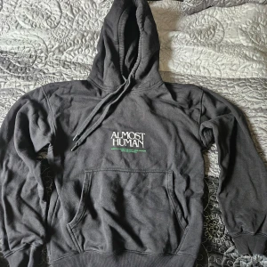 Svart hoodie från Vailent - Säljer en cool svart hoodie från Vailent med texten 'Almost Human' tryckt på både fram- och baksidan. Den har en stor grön ansiktsgrafik på baksidan och en känguruficka framtill. Perfekt för höst och vinter, den är både bekväm och stilren. Passar perfekt för en avslappnad look!