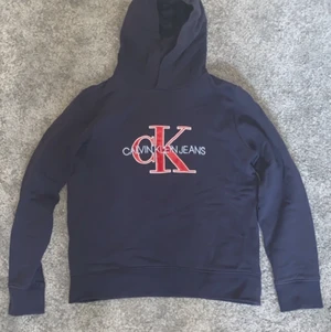 Calvin Klein hoodie storlek 164 - En fin marinblå Calvin Klein hoodie med in broderat Calvin Klein jeans i ljusblått och tryckt CK i rött Trycket är lite slitet efter tvätt men ändå i bra skick (8/10)